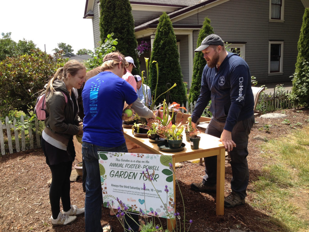 People in a yard next to a house, standing at a table with potted plants on top, with a sign in front reading, "Annual Foster-Powell Garden Tour"